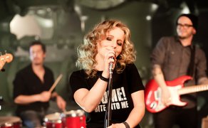 Band mit Sängerin, Schlagzeuger und Gitarrist bei Live-Auftritt, Sängerin hält Mikrofon und trägt T-Shirt mit Aufdruck.