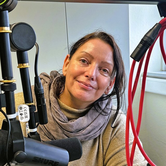 Frau mit braunem Schal sitzt vor einem schwarzen Mikrofon, im Hintergrund Fenster und rote Kabel an der Wand.