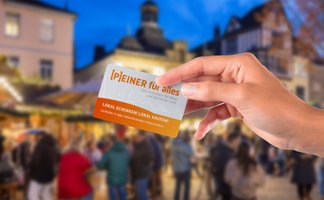 Hand hält eine Karte mit dem Text „Peiner für alles“ und „Lokal schenken, lokal kaufen!“ vor unscharfem Weihnachtsmarkt mit Menschen und Gebäuden.