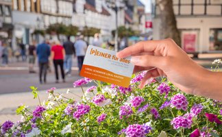 Hand hält den Stadtgutschein „Peiner für alles – Lokal schenken, lokal kaufen“ vor einer belebten Innenstadt mit Blumen und Passanten im Hintergrund.