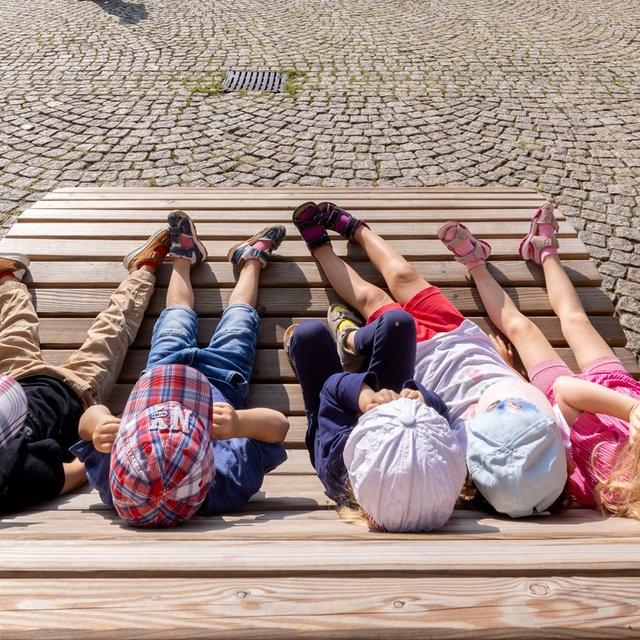 Das Bild zeigt vier Kinder, die auf eine Holzliege auf dem Historischen Marktplatz in Peine liegen. 