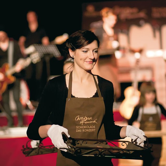 Frau mit Schürze und weißen Handschuhen trägt Tablett mit Schokoladenstücken vor einer Bühne mit Musikern.