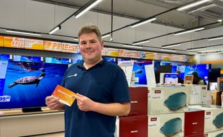 Ein Mitarbeiter steht in einem Elektrofachmarkt und hält einen „[P]EINER für alles“-Gutschein in der Hand. Im Hintergrund sind Fernseher und weitere Elektrogeräte ausgestellt.
