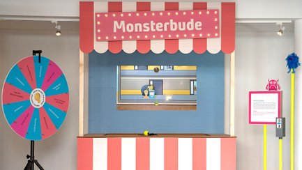 Wurfbude mit rot-weiß gestreiftem Baldachin und Schild „Monsterbude“, links ein Glücksrad, rechts ein Infotext und Monster-Figuren auf einem Ständer.