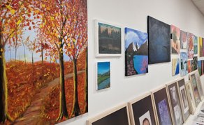 Menschenmenge in einer Galerie mit zahlreichen farbigen Gemälden an den Wänden, darunter ein großes Bild einer sitzenden Figur in Grün auf gelbem Hintergrund.