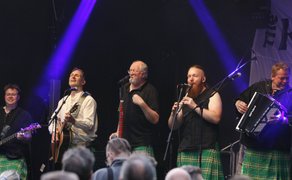 Live-Konzert beim Highland Gathering in Peine: Folk-Band in Kilts steht auf der Bühne unter violettem Bühnenlicht und begeistert das Publikum mit schottischer Musik, Gesang, Gitarre, Flöte und Akkordeon.