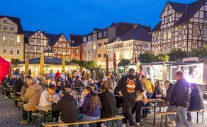 Menschen sitzen und stehen an Tischen auf einem belebten Platz mit Fachwerkhäusern und beleuchteten Verkaufsständen.