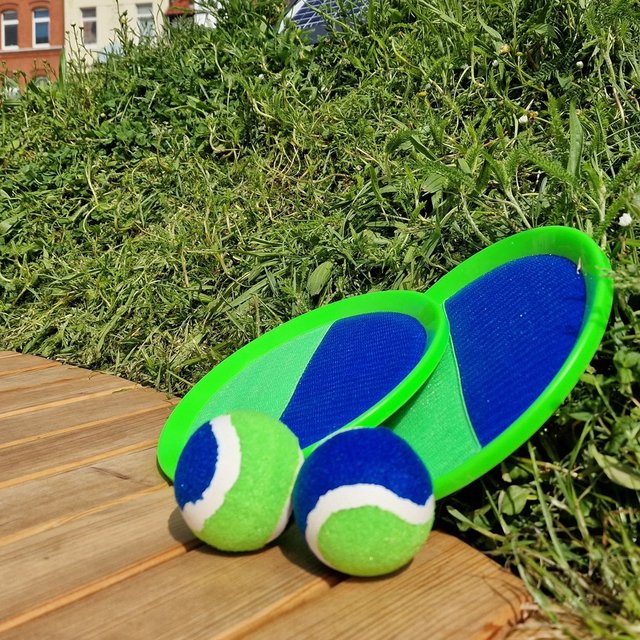 Zwei grün-blaue Wurfspiele mit passenden Bällen liegen auf einer Holzplattform vor einer bepflanzten Grünfläche; im Hintergrund sind Fachwerkhäuser und Gebäude der Peiner Innenstadt zu erkennen.