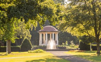 Der Pavillon im Peiner Stadtpark steht umgeben von großen Bäumen am Ende eines sonnigen Weges.