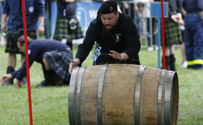 Teilnehmer in schottischer Tracht mit Kilt und Barett rollt konzentriert ein Holzfass über die Wiese bei einem traditionellen Wettkampf des Highland Gatherings, während weitere Teilnehmer und Zuschauer im Hintergrund zu sehen sind.