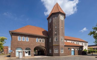 Ein großes Backsteingebäude mit rotem Ziegeldach und markantem Uhrturm steht an einem Platz. Der Turm hat auf mehreren Seiten Zifferblätter. Das Gebäude verfügt über Rundbögen im Eingangsbereich und mehrere Fensterreihen. Im Vordergrund ist eine gepflasterte Fläche zu sehen, rechts und links stehen einzelne Bäume. Der Himmel ist blau mit wenigen Wolken.