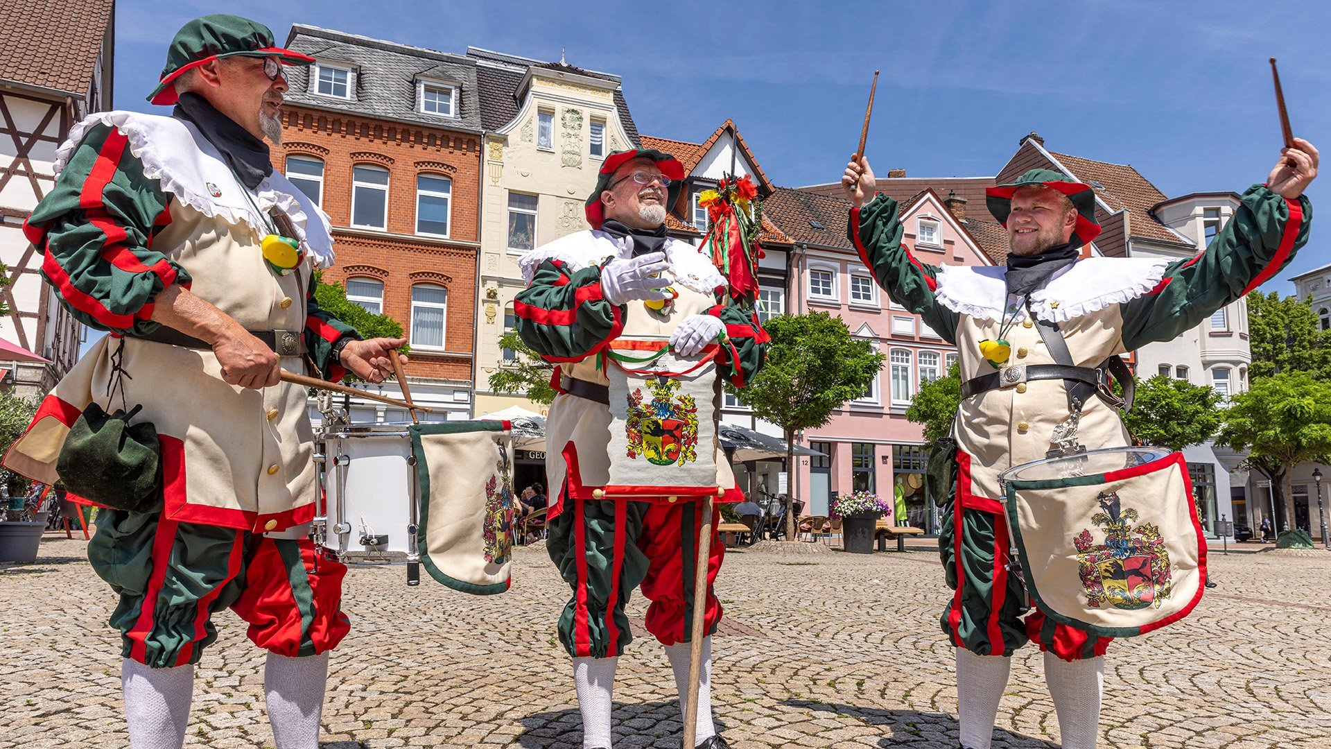Drei Männer in historischen, grün-roten Uniformen mit Trommeln stehen auf einem gepflasterten Marktplatz in Peine und spielen fröhlich Musik vor bunten Altstadthäusern bei sonnigem Wetter.