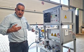 Ein zeigt auf technische Geräte auf einem Gestell und spricht in die Kamera.