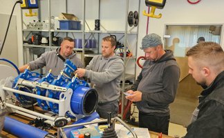Vier Männer stehen in einer Werkstatt an einer technischen Apparatur, in die ein Kunststoffrohr eingespannt ist.
