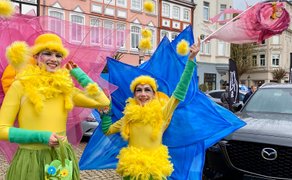 Zwei Personen in gelben Kostümen mit grünen Ärmeln und großen bunten Flügeln stehen auf einem historischen Marktplatz neben einem schwarzen Auto.