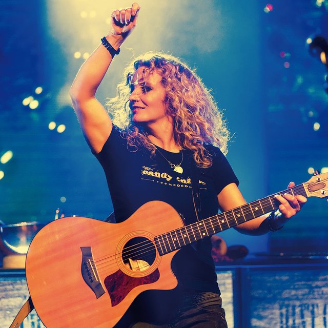 Das Bild zeigt eine Frau auf einer Bühne mit einer Gitarre in der Hand. Sie hebt den rechten Arm in die Luft.