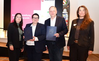 Vier Personen stehen bei der Übergabe von Förderbescheiden des Programms „Zukunftsräume Niedersachsen“ vor einem Bildschirm mit Präsentationsfolie in einem historischen Saal.