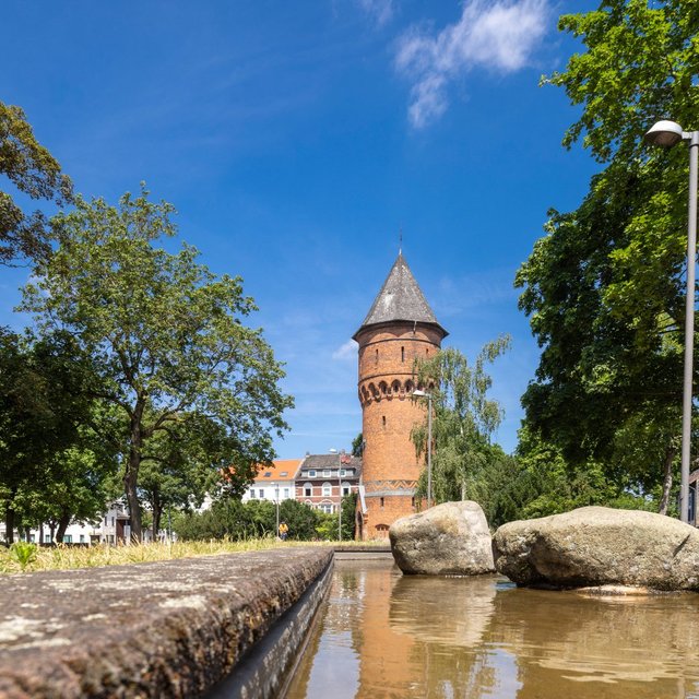 Im Vordergrund verläuft ein flacher Wasserlauf mit Steinen, dahinter erhebt sich ein runder Backsteinturm mit spitzem Dach. Der Turm ist von Bäumen und Gebäuden umgeben. Die Szene ist bei sonnigem Wetter aufgenommen, der Himmel ist blau. Die Perspektive ist niedrig und lenkt den Blick entlang des Wassers auf den historischen Turm.