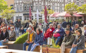 Zahlreiche Menschen sitzen und stehen auf dem Historischen Marktplatz in Peine, viele auf Holzbänken zwischen bepflanzten Elementen, und verfolgen entspannt ein kulturelles Programm in lebendiger Innenstadtatmosphäre mit Fachwerkhäusern und Cafés im Hintergrund.