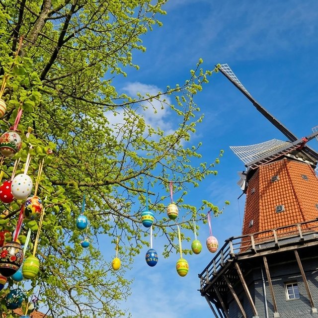 Windmühle mit rot gedecktem Kegel und hölzernem umlaufenden Balkon neben Baumzweigen mit zahlreichen bunt bemalten Ostereiern an Bändern.
