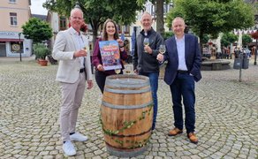 Vier Personen stehen beim Peiner Weinfest mit Weingläsern um ein Holzfass auf einem Marktplatz.