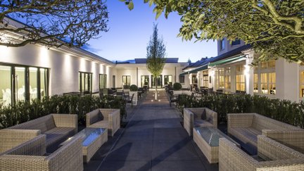 Beleuchteter Innenhof eines modernen Hotels in der Abenddämmerung mit Sitzgruppen aus Rattan, gepflastertem Weg, Bäumen und hell erleuchteten Gebäudefassaden auf beiden Seiten.