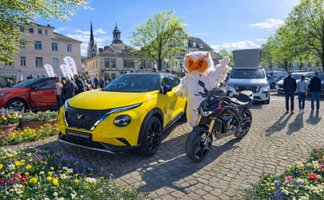 Gelbes Auto und schwarzes Motorrad auf gepflastertem Platz mit Person im Eulen-Kostüm und mehreren Besuchern bei sonnigem Wetter.