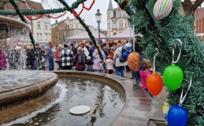 Ein geschmückter Brunnen auf einem Marktplatz, dekoriert mit grünen Girlanden und bunten Ostereiern im Vordergrund. Im Hintergrund stehen viele Menschen, darunter Familien mit Kindern, vor Ständen und Zelten und nehmen an einer Veranstaltung teil. Historische Gebäude umgeben den Platz.