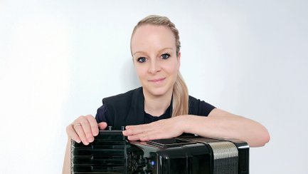Die Musikerin blickt lächelnd in die Kamera und lehnt mit den Armen auf ihrem schwarzen Digital-Akkordeon. Sie steht vor einem hellen, neutralen Hintergrund und trägt ihre blonden Haare zu einem seitlichen Zopf gebunden.