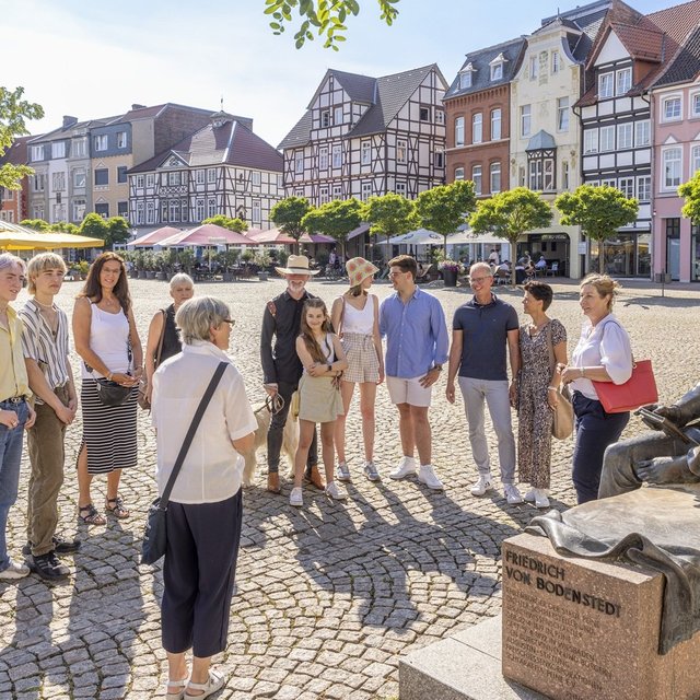 Eine Gruppe von Menschen steht bei einer Stadtführung auf dem historischen Marktplatz neben der Friedrich-von-Bodenstedt-Statue vor Fachwerkhäusern.