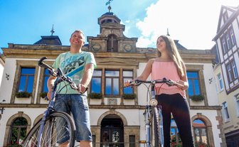 Zwei Personen mit Fahrrädern stehen vor einem historischen Gebäude mit großen Fenstern und einem Uhrenturm.