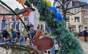Osterbrunnen mit hängenden Dekorationen, darunter ein brauner Hasenanhänger, auf einem belebten Marktplatz mit Menschen und Gebäuden im Hintergrund.