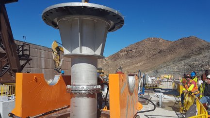 Große zylindrische Rohrleitung mit orangen Schutzvorrichtungen auf einer Baustelle in einer bergigen Wüstenlandschaft mit mehreren Personen in Schutzkleidung und Helmen