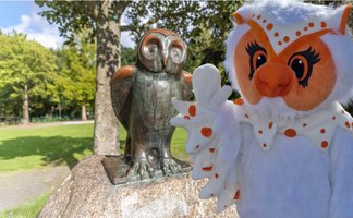 Uhlinchen steht neben einer Eulenstatue im Grünen in Peine und winkt freundlich in die Kamera.