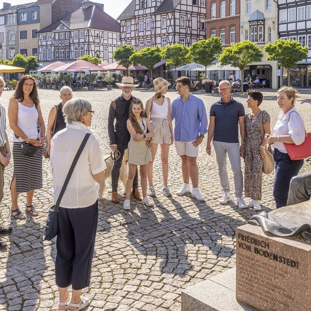 Eine Gruppe von Menschen steht bei einer Stadtführung auf dem historischen Marktplatz neben der Friedrich-von-Bodenstedt-Statue vor Fachwerkhäusern.