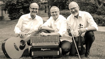 Drei Musiker der Gruppe Wenos sitzen lachend auf einer Wiese im Freien und posieren gemeinsam mit ihren Instrumenten für ein schwarz-weißes Gruppenfoto. Zu sehen sind eine Gitarre, ein Akkordeon und eine Geige. Alle tragen helle Hemden, im Hintergrund befindet sich eine unscharfe Garten- oder Parklandschaft.