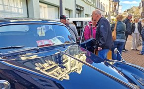 Oldtimer mit glänzender Motorhaube, auf der sich ein historisches Gebäude spiegelt, umgeben von Menschen auf einer gepflasterten Straße..