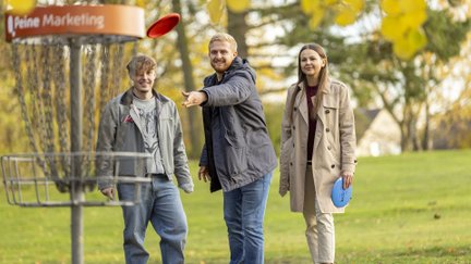 Drei junge Erwachsene spielen Discgolf an einem Korb im Grünen in Peine, Freizeitaktivität im Park bei herbstlicher Stimmung.