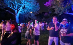 Silent Disco in Peine bei Nacht: Feiernde Menschen tanzen im Park mit farbig leuchtenden Funkkopfhörern zwischen blau und violett angestrahlten Bäumen, lockere Sommerstimmung mit Musik, Getränken und Lichteffekten.