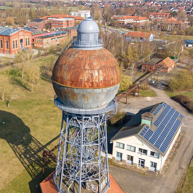 Luftaufnahme eines alten Kugelwasserturms auf einem Industriegelände mit angrenzendem Gebäude und weiteren Industrieanlagen im Hintergrund