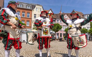 Drei Männer in historischen Uniformen des Peiner Freischießens spielen auf Trommeln auf einem Marktplatz.