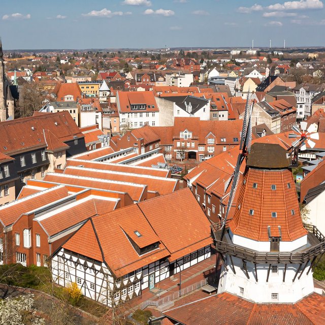 Luftaufnahme der Peiner Innenstadt mit rotem Ziegeldächern, historischer Windmühle und Kirchturm, dicht bebauten Straßenzügen und weiter Landschaft am Horizont bei sonnigem Wetter.
