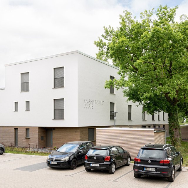 Modernes Wohngebäude mit weißer Fassade und braunem Sockel, davor mehrere geparkte Autos und ein großer Baum.