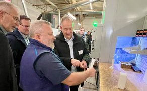 Mehrere Männer, einer von ihnen in Arbeitskleidung, stehen an einer Werkbank mit Kunststoffblöcken in einer Industriehalle.