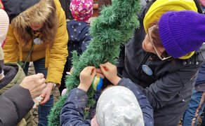 Mehrere Personen, darunter ein Kind mit grauer Mütze und ein Erwachsener mit lila Mütze, schmücken gemeinsam einen grünen Osterbrunnenkranz mit bunten Ostereiern.