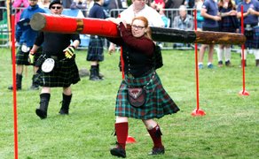 Frau in traditionellem Kilt trägt Baumstamm bei einem Baumstammslalom-Wettbewerb auf einer Wiese mit Zuschauern im Hintergrund.