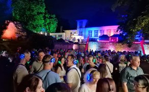 Besucher mit leuchtenden Funk-Kopfhörern feiern bei der Silent Music Party im Peiner Burgpark vor farbig illuminierter Kulisse und DJ-Bühne im Sommerabend.