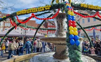 Der Osterbrunnen auf dem Peiner Marktplatz ist mit grünen Girlanden und bunten Ostereiern geschmückt, während sich viele Menschen rund um den Brunnen versammeln.