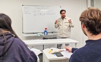 Ein Mann steht vor einem Whiteboard mit Formeln und spricht in Richtung von zwei Personen, die sich Notizen machen.
