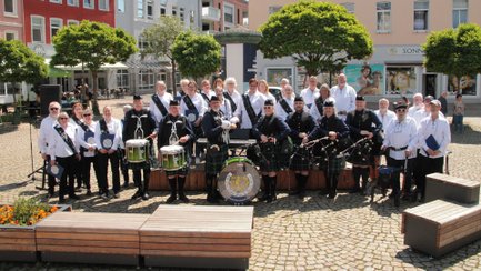 Eine große Gruppe der Scottish Folk Singers und Mitglieder des Scottish Culture Club Peine steht gemeinsam auf dem Historischen Marktplatz. Die Sängerinnen und Sänger tragen überwiegend weiße Oberteile, während einige Musiker in traditioneller schottischer Kleidung mit Dudelsäcken und Trommeln vor der Gruppe positioniert sind. Im Hintergrund sind Bäume und Gebäude der Innenstadt zu sehen.